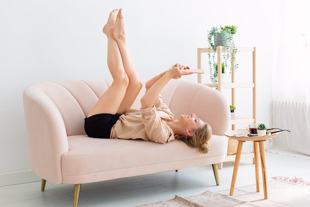 Happy woman lies on her back on a beige sofa legs up looks at a digital tablet