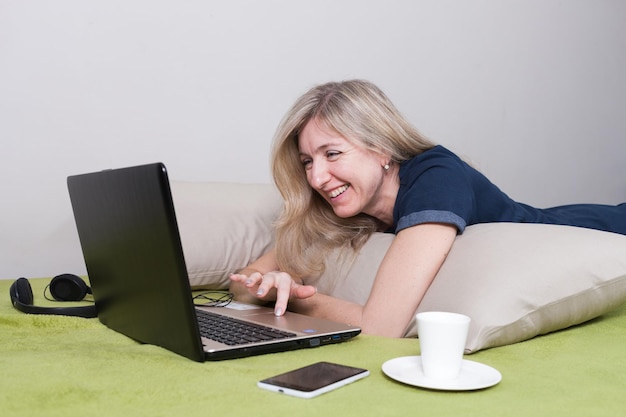 Happy woman lies on stomach on pillows on green sofa looks at laptop and smiles