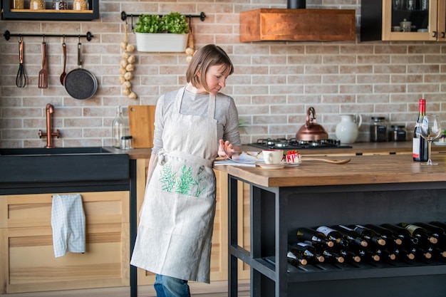 Happy woman in a linen apron standing in the kitchen at the table examines a magazine.