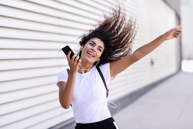 happy woman listening music while dancing by wall in city