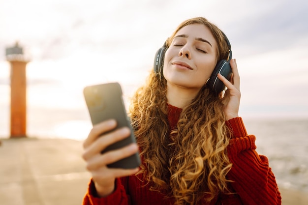 Happy woman listening nice music by the waterfront while wearing headphones by a lighthouse