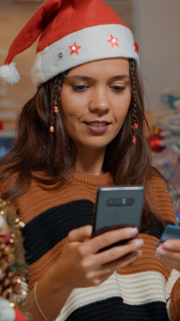 Happy woman looking for christmas gifts at decorated home. Young adult wearing santa hat using smartphone and credit card to shop online for seasonal festive celebration dinner party