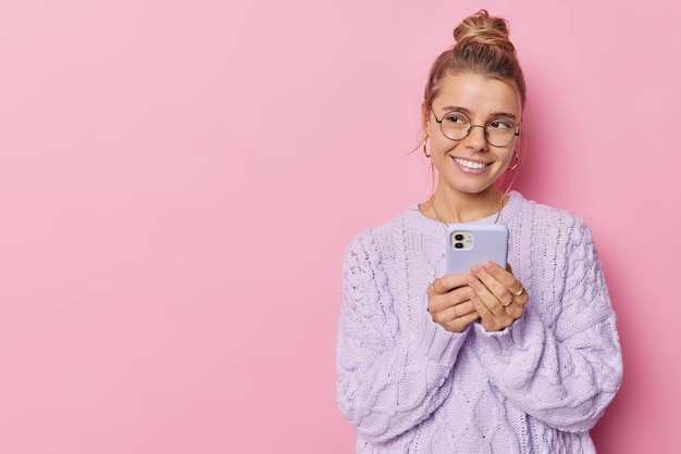 Happy woman looks with glad thoughtful expression away chats on\
mobile messanger uses smartphone application wears round spectacles\
knitted jumper isolated over pink background blank space on\
left