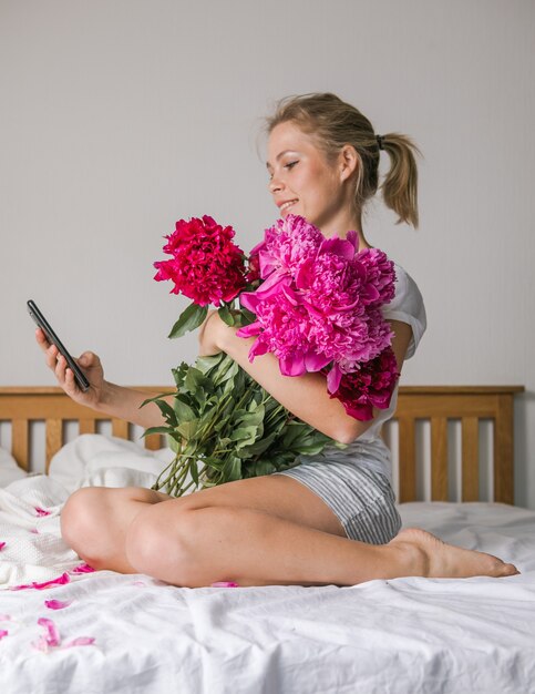 Happy woman lying on the bed wearing pajamas, enjoying tulip flowers bouquet of peony and using phone