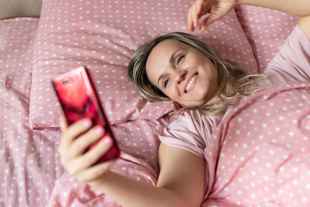 Счастливая женщина лежит в постели со смартфоном в руках Просыпается перед сном Проблемы со сном Молодая женщина использует смс-сообщения на смартфоне или делает селфи на кровати дома