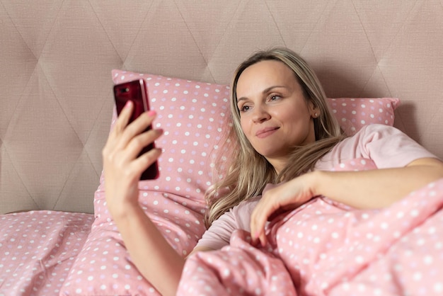 Donna felice sdraiata a letto con lo smartphone in mano svegliarsi andando a letto problemi di sonno giovane donna che usa la messaggistica di testo dello smartphone o si fa un selfie sul letto a casa