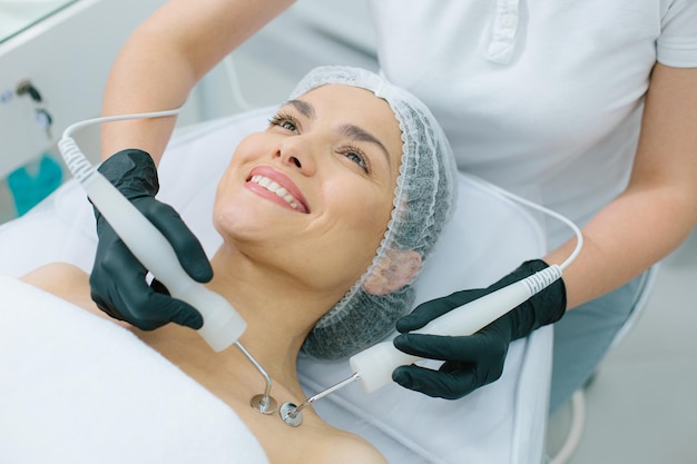 Happy woman lying and smiling and a beautician touching her collarbone with two metal prongs
