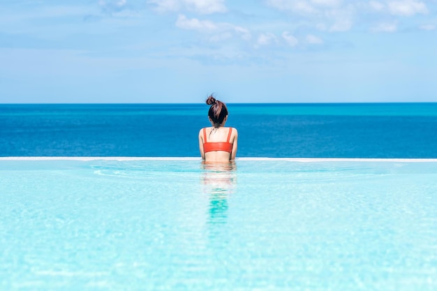 Happy woman in orange swimsuit swimming in infinity pool at luxury hotel against ocean front. young female enjoy in tropical resort. Relaxing, summer,  travel, holiday, vacation and weekend concept