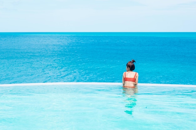 Happy woman in orange swimsuit swimming in infinity pool at luxury hotel against ocean front young female enjoy in tropical resort Relaxing summer travel holiday vacation and weekend concept