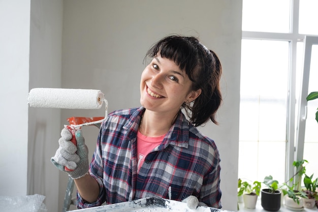 Photo happy woman in paint roller and white paint for walls in hands closeup portrait construction work and cosmetic repairs in house wall painting tinting finishing work with your own hands