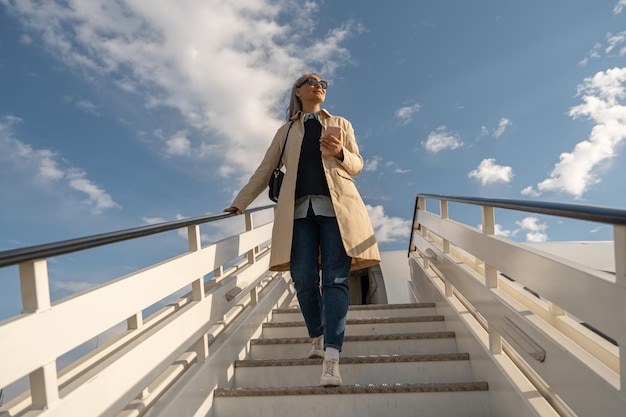 Счастливая женщина-пассажирка в солнцезащитных очках спускается по лестнице самолета после приземления в аэропорту