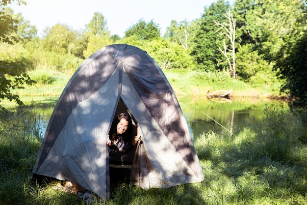 체크 무늬 셔츠를 입은 행복한 여성이 아침에 강둑에서 하이킹을 하며 관광 텐트 밖을 바라보고 있습니다. 야생 가족 휴가와 모험에서 밤새 자연에서 캠핑
