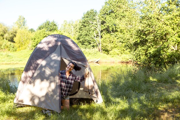 체크 무늬 셔츠를 입은 행복한 여성이 아침에 강둑에서 하이킹을 하며 관광 텐트 밖을 바라보고 있습니다. 야생 가족 휴가와 모험에서 밤새 자연에서 캠핑