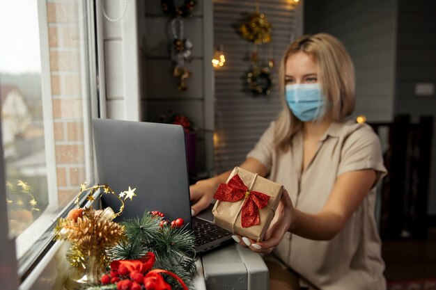 Happy woman in protective mask, taking gift in hand and doing online shopping at home to buy christmas present during quarantine. Focused on gift.