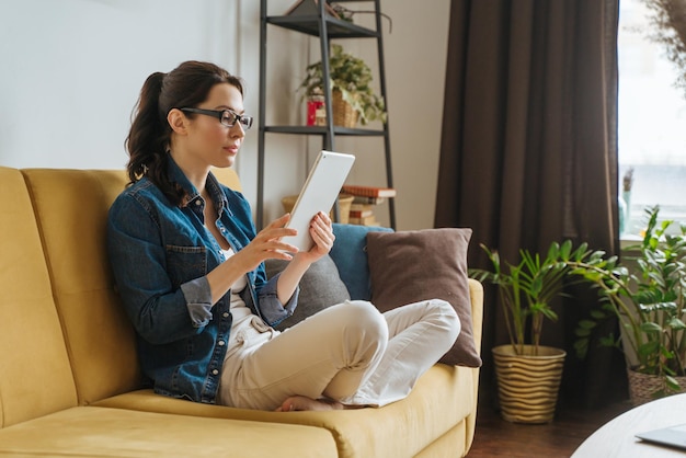 Счастливая женщина, читающая книгу в читалке электронных книг, сидя на диване у себя дома