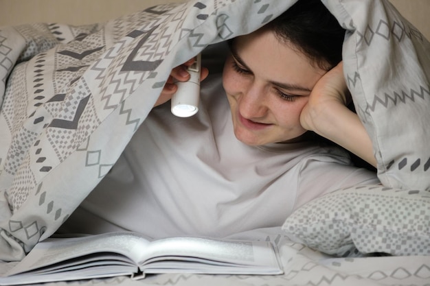 A happy woman reads a book lying under a blanket