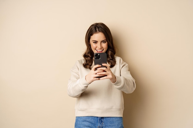 Happy woman recording video, shooting photo on smartphone camera and smiling, standing over beige background