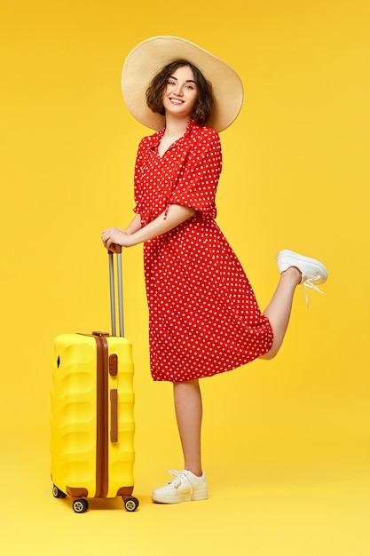 Happy woman in red dress with suitcase going traveling on yellow background.