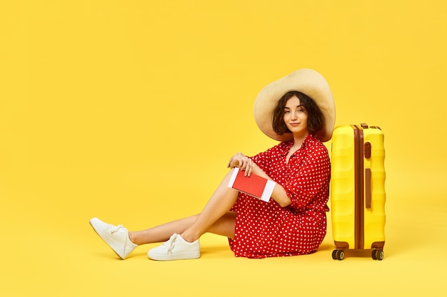 Happy woman in red dress with suitcase and passport going traveling on yellow background.