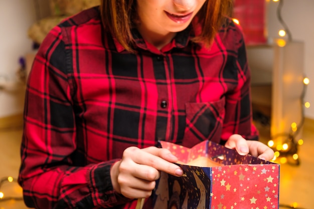 빨간 셔츠를 입은 행복한 여성은 생일 안에 빛이 들어 있는 패키지에서 크리스마스 선물을 엽니다.