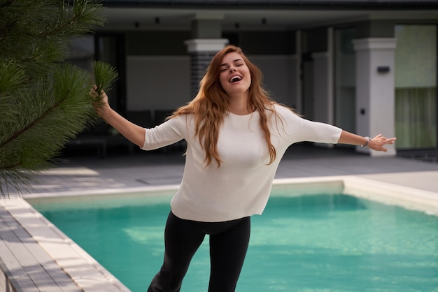 Happy woman rejoicing while spending time near her swimming pool at the modern villa. Ginger woman feeling great at the morning