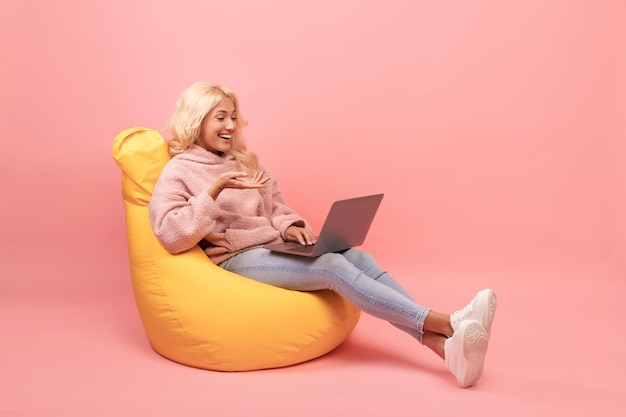 현대적인 가제트 분홍색 배경을 사용하여 노트북 여학생과 함께 빈백 의자에서 휴식을 취하는 행복한 여성
