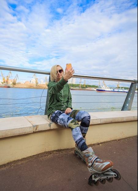 Happy woman in roller skates sitting and making selfie photo on smart phone on riverside