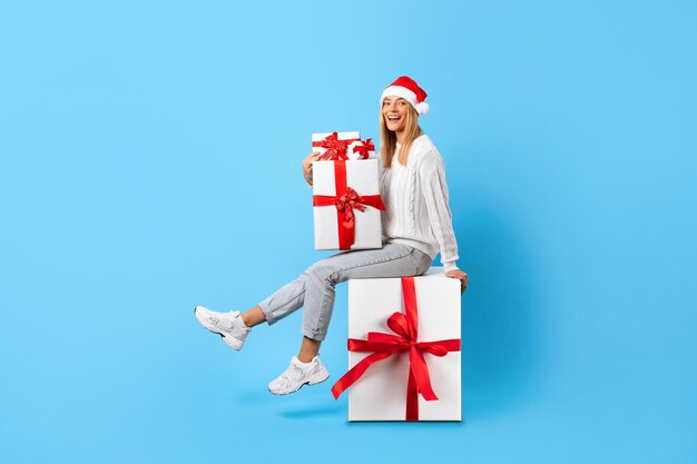 Happy woman in santa hat with presents sitting on huge gift box on blue studio background