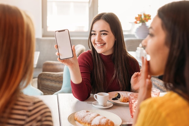 Happy woman showing her friends information on smartphone screen while sitting in cafe