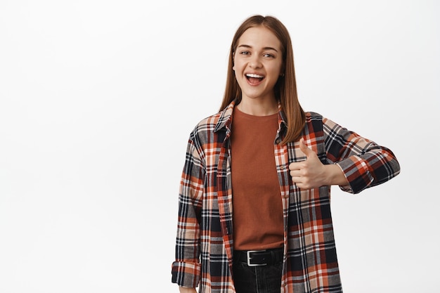 Happy woman shows thumbs up and smiles, stands against white wall