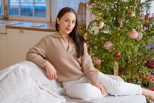 Фото Счастливая женщина сидит на диване в свитере рядом с естественным рождественским деревом праздничного настроения