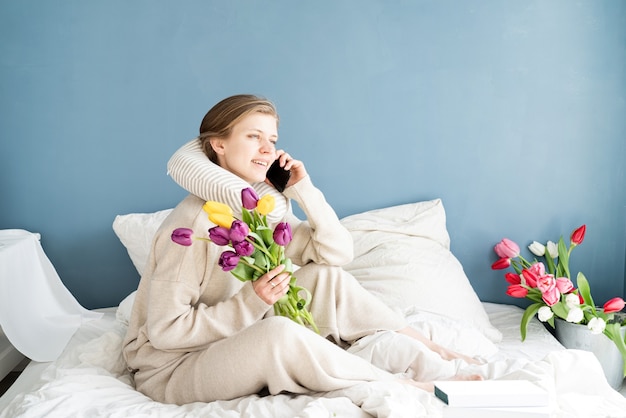 Happy woman sitting on the bed wearing pajamas talking on the phone, blue wall background