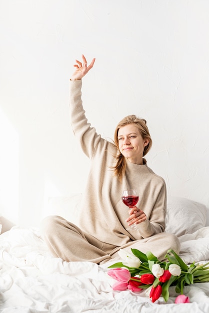 Felice donna seduta sul letto in pigiama, con piacere godersi i fiori e un bicchiere di vino rosso