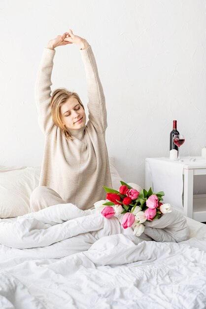 Happy woman sitting on the bed wearing pajamas, with pleasure enjoying flowers and stretching