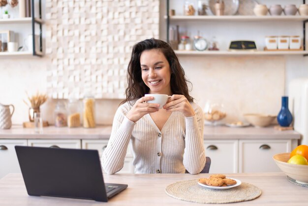 Happy woman sitting in front of laptop at kitchen table watching movie on computer and drinking