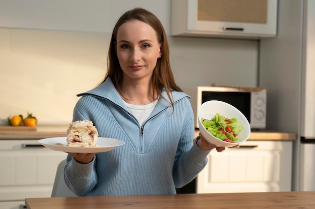Счастливая женщина, сидящая за кухонным столом, выбирает между тортом и овощным салатом.