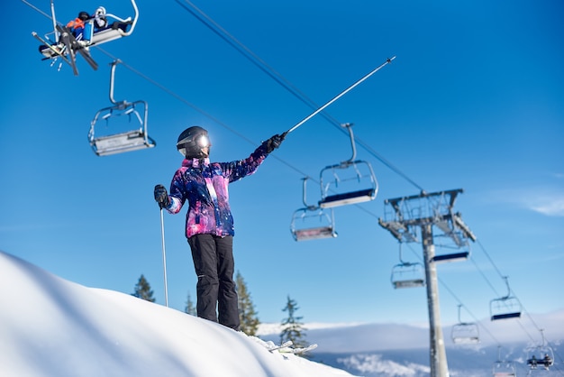 Happy woman skier standing near ski lift on snow-covered mountain slope. Sunny day during winter vacation. General view.