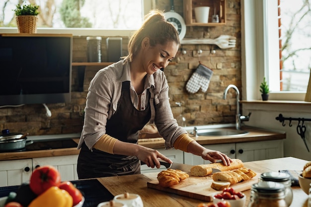 Happy woman slicing bread with a knife while preparing food in the kitchen