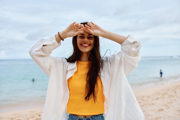 Happy woman smile with teeth with long hair brunette walks along the beach in a yellow tshirt denim shorts and a white shirt near the sea summer journey and feeling of freedom balance