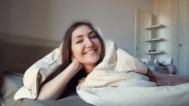 Happy woman smiles and looks at camera from under soft blanket on large bed in apartment close-up