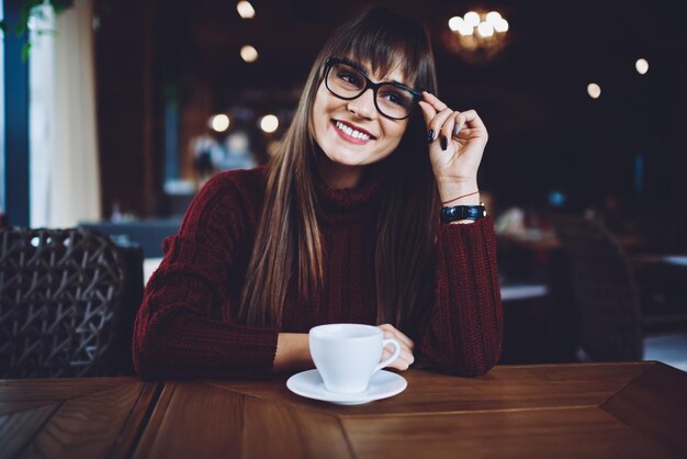 カフェイン飲料とカフェテリアのテーブルに座って視力矯正のための眼鏡で幸せな女性