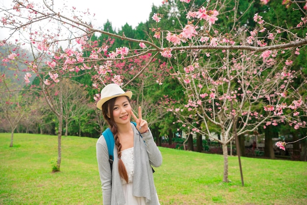 happy woman standing in front of cherry tree and showing victory gestures taking picture happy enjoying sakura flowers bloom in japan vacation trip.