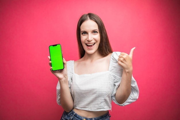 Happy woman standing and hold smartphone and thumbs up on pink background. Green screen phone screen