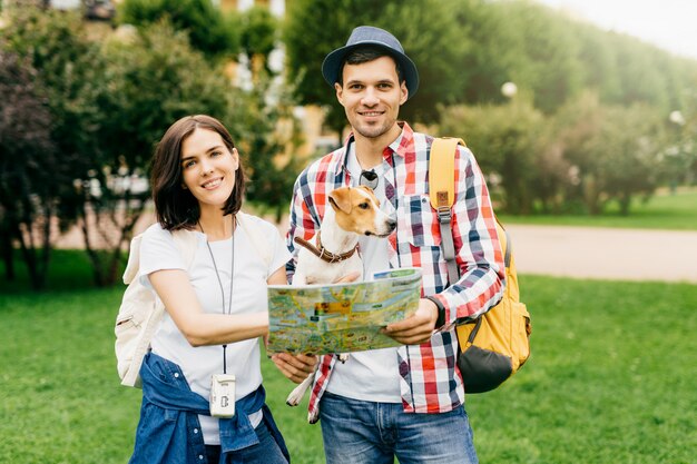 Happy woman standing near her husband and dog, having together walk across unknown city, looking in map while looking for different places of interest. Couple of young travelers standing outdoors