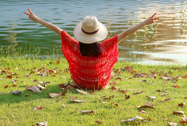 Happy Woman in Straw Hat Raising Arm with V Sign Sitting on the Lakeside