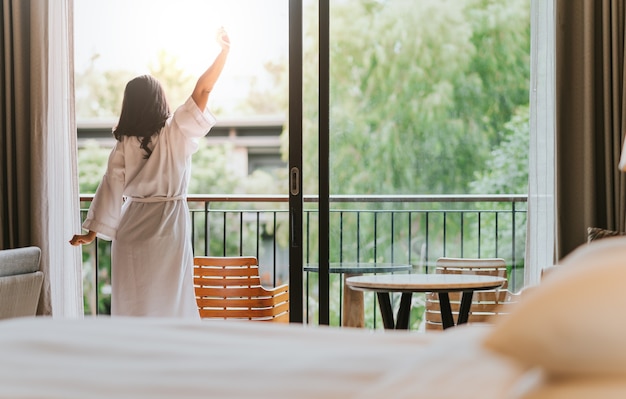 Happy woman stretches and opens the curtains at window in morning.