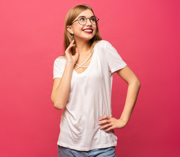 Happy woman in t-shirt posing with arm on hip while having idea and looking at the camera over pink background.