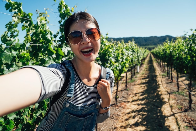 Turista donna felice che cammina nel campo di uva soleggiato prendendo selfie con alberi e piante. giovane ragazza sorridente allegramente prendendo autoritratto sotto la luce del sole in cantina. il viaggiatore visita la valle di napa in primavera