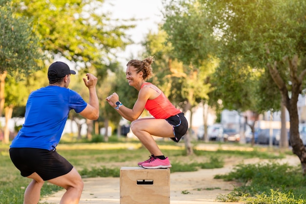 Счастливая тренировка женщины в парке рядом со своим тренером по фитнесу. Она использует прыжковый ящик.