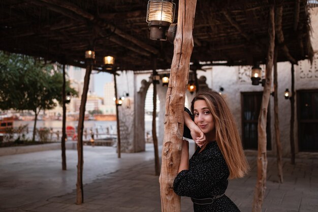 Happy woman traveler wearing black dress walking through the streets of an old Arab town or village in the middle of the desert. Traditional Arabic oil lamps in Al Seef Dubai street